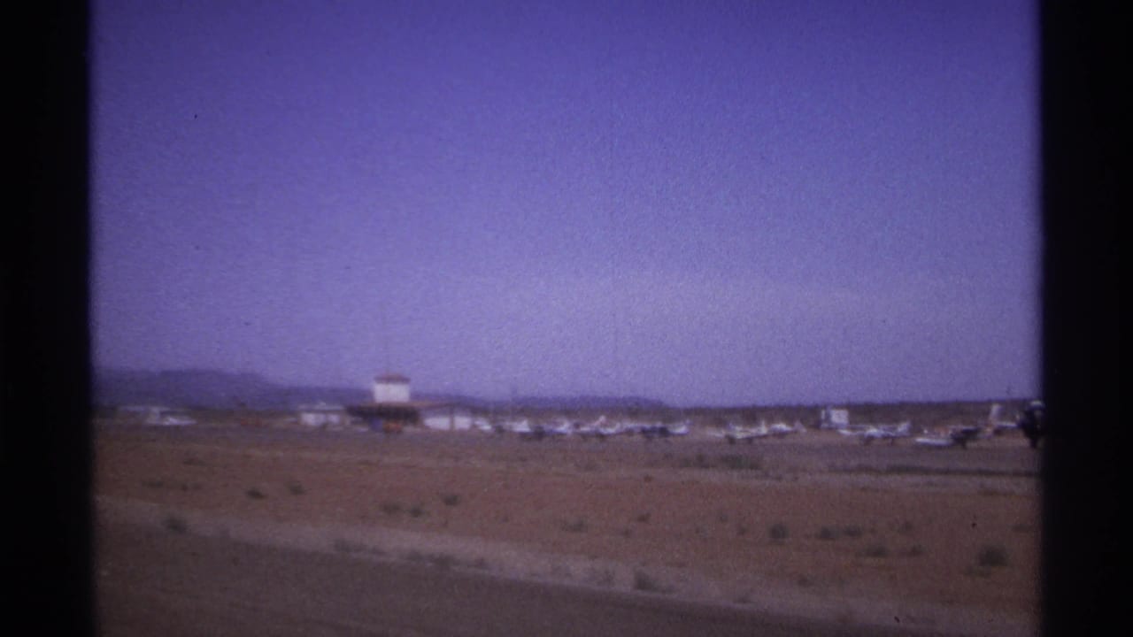 Wide view of a desert airfield with small aircraft and a tower-like building beyond the runway.