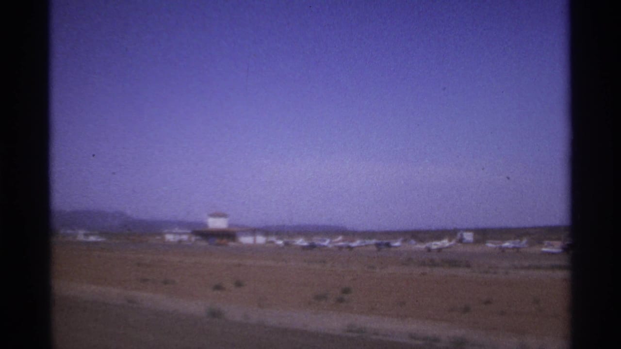 Distant airport runway area with parked small aircraft, low buildings, and dark film borders.