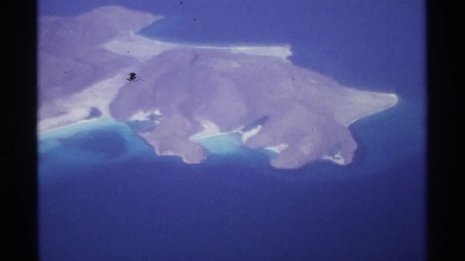 1974 La Paz travel footage shifts to a high aerial view of a coastal island with rocky pale terrain, turquoise shallows, and dark blue sea.