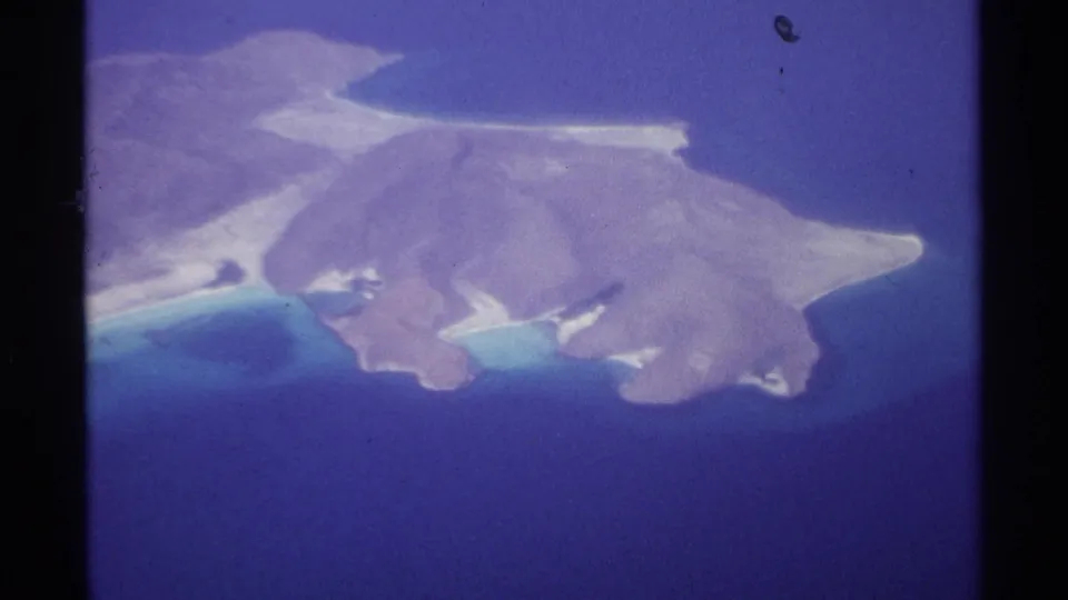 Final 8mm frame showing a coastal island near La Paz from above, with sandy or rocky points, bright shallows, and saturated blue water.