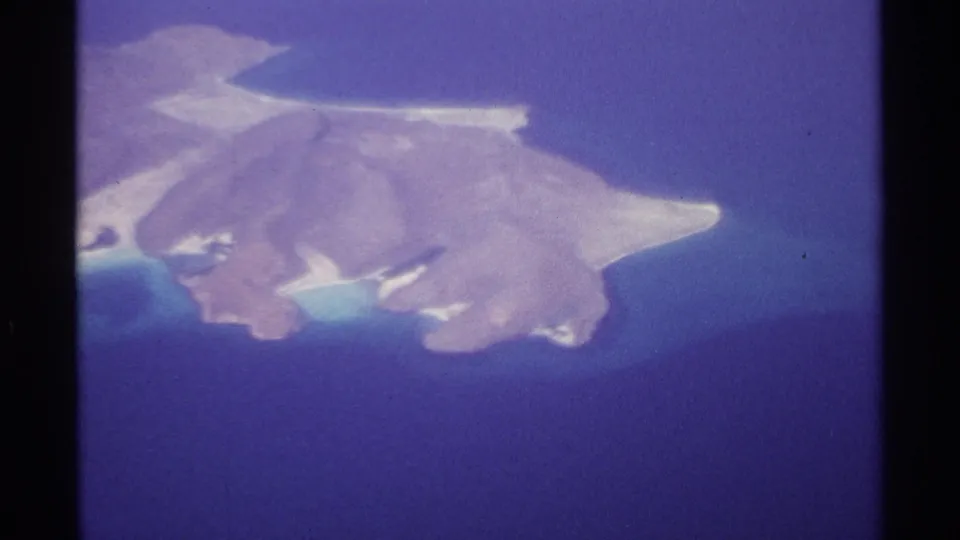 Vintage 8mm aerial view near La Paz showing the island closer in frame, with sandy points, rocky edges, and clear blue water.