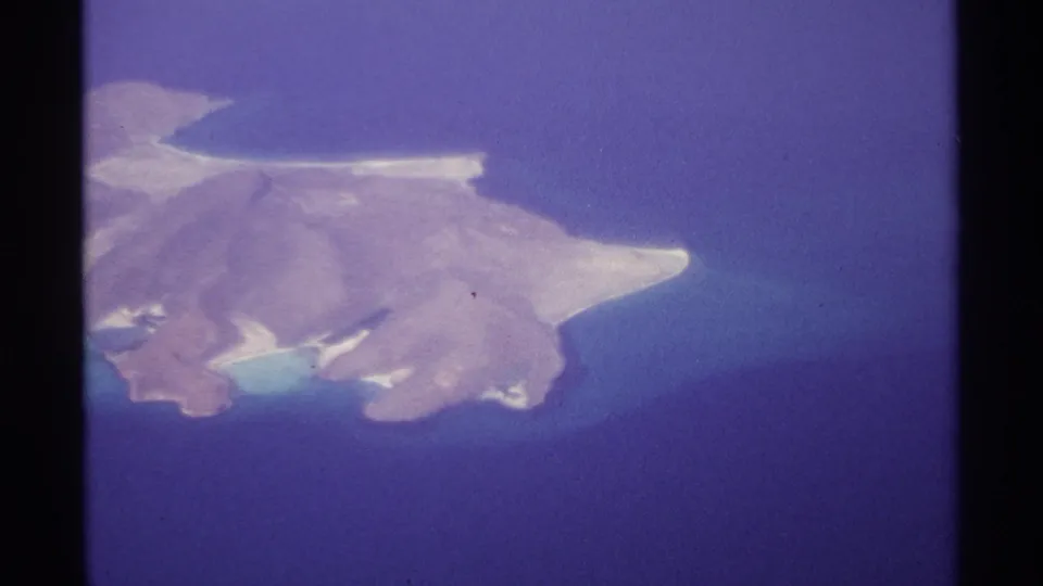 1974 home-movie aerial frame near La Paz, Mexico, showing the brown island set against deep blue water and lighter coastal shallows.