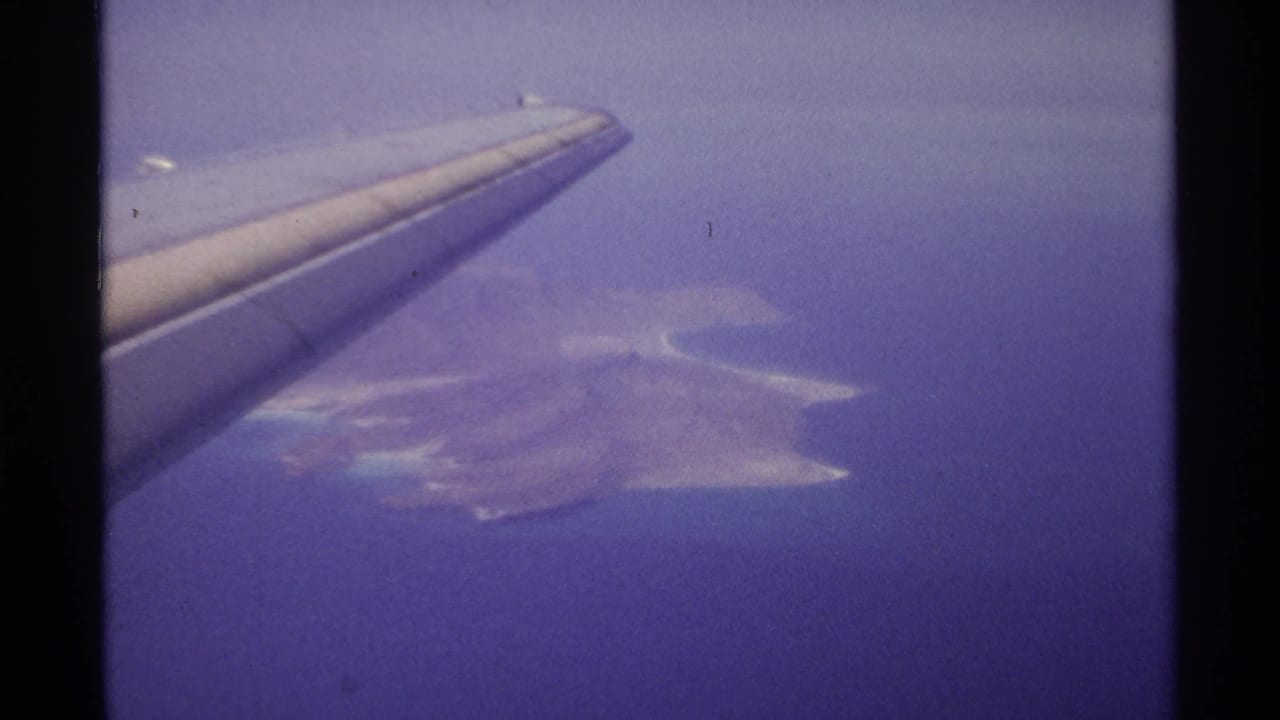 Airplane wing crosses the frame above an island and surrounding ocean.