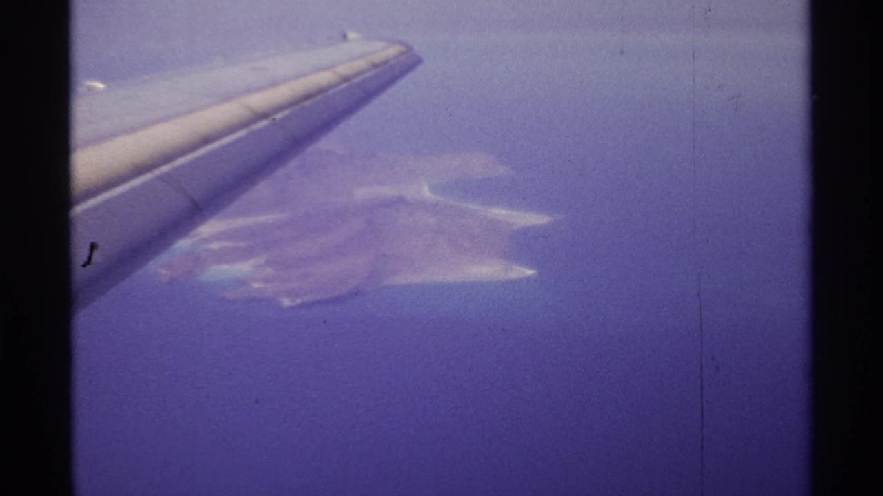 1974 8mm travel frame from the La Paz, Mexico area showing an airplane wing over hazy blue water and pale coastal shapes.