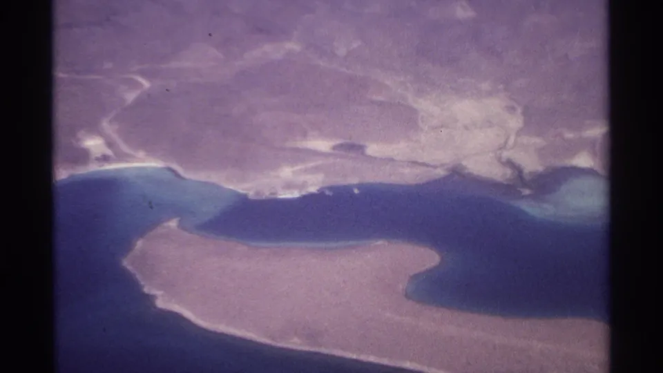 1974 aerial home-movie frame showing a sandy island and irregular coastline near La Paz, Mexico, with blue water between them.