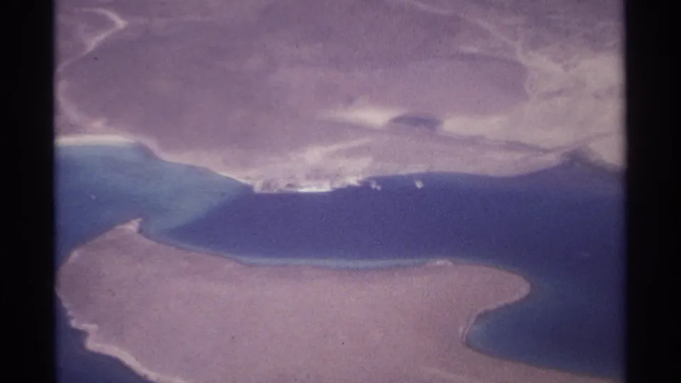 1974 8mm aerial frame near La Paz, Mexico, showing blue coastal water, sandy land and a distant white shoreline structure.