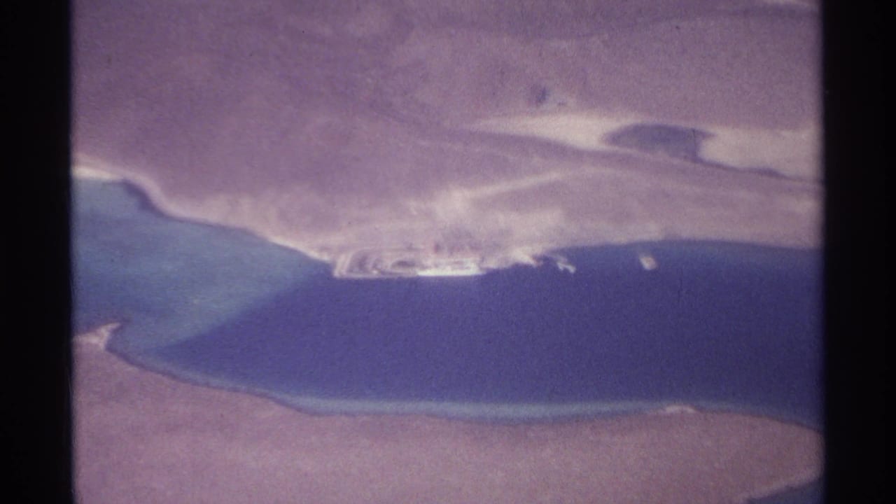 Aerial coastline with blue water, sandy shore and a small pale structure near the waterline.