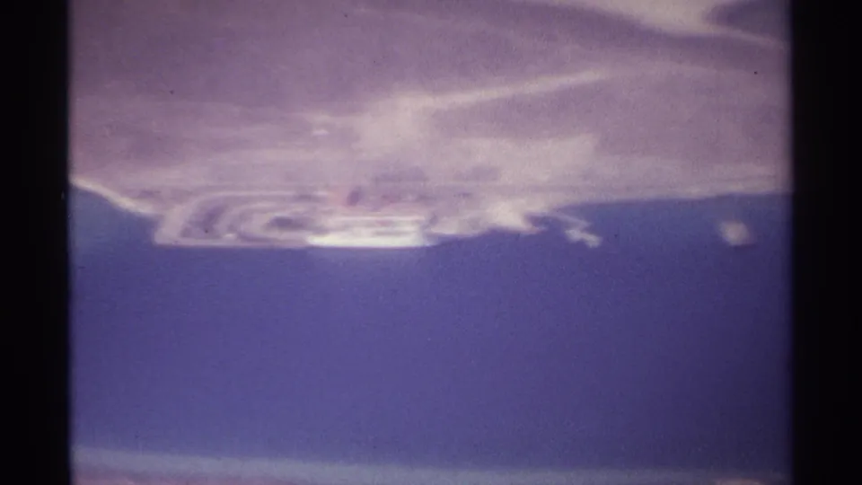 1974 8mm aerial frame near La Paz with blue water below, pale coastal land above and a bright distant shoreline feature.
