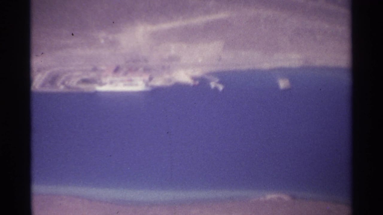 1974 8mm aerial view over La Paz, Mexico, showing blue coastal water, pale shoreline development, and road traces near the waterfront.