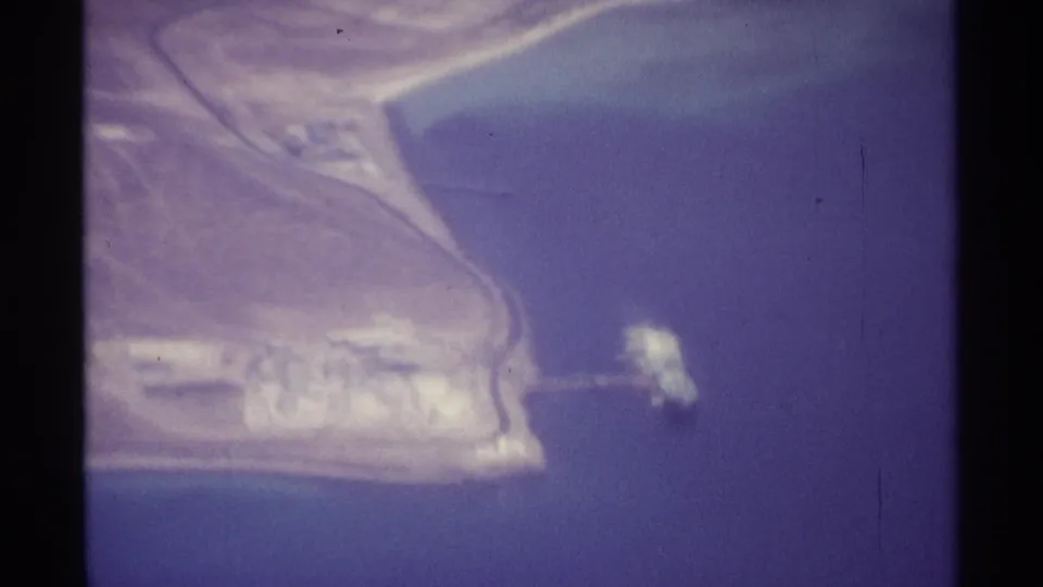 1974 La Paz aerial view showing shoreline buildings and roads beside blue water, with a small pale offshore object near the coast.
