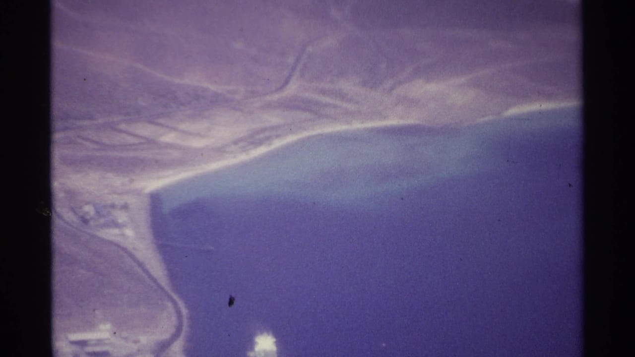 Aerial coastline with dry ridges, roads, deep blue water, and a bright object near shore.
