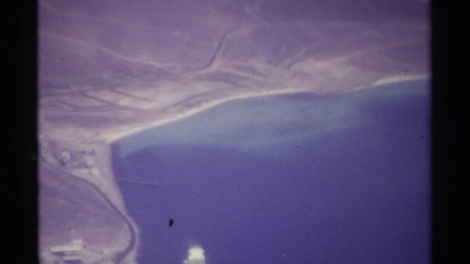 Aerial 8mm frame near La Paz, Mexico, with arid coastal land, curving shoreline, roads, ocean, and a bright object on the water.
