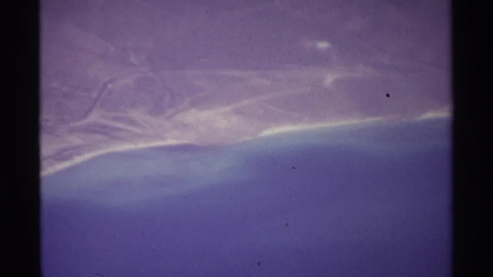 1974 8mm aerial frame near La Paz, Mexico, showing hazy dry coastal terrain, a pale shoreline, and blue ocean below.