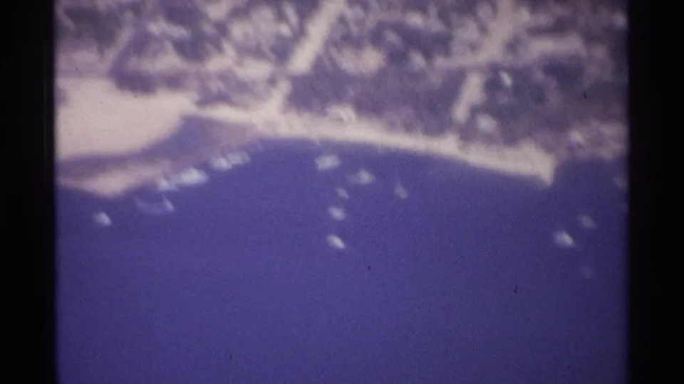 1974 La Paz aerial frame with curved coastline, clustered small boats, pale beach, trees, and low-rise settlement near the water.