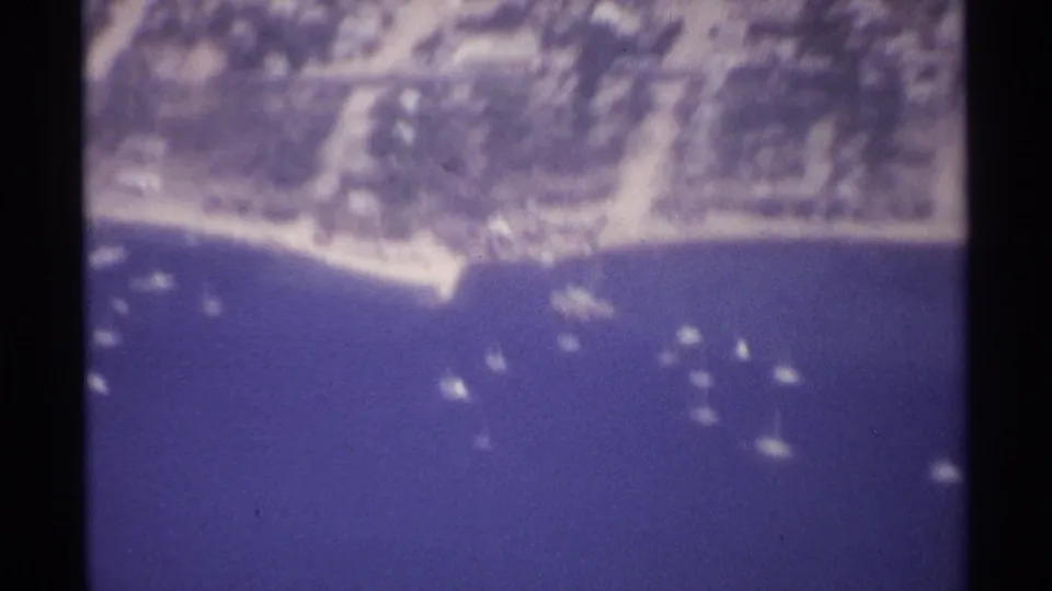 1974 8mm aerial view of La Paz's waterfront area with low-rise homes, trees, a sandy edge, and numerous small boats on the water.