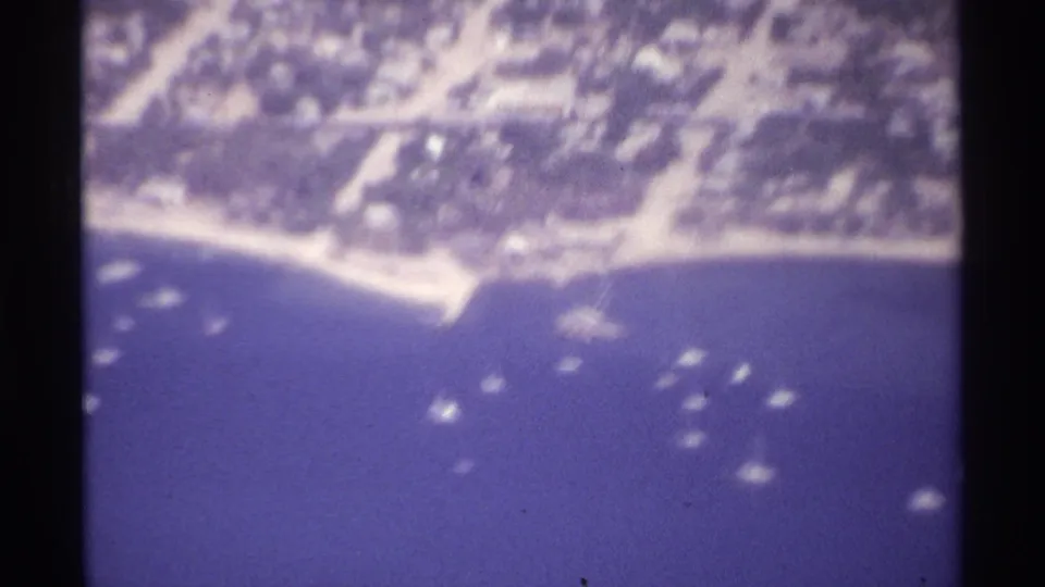 Late frame from the 1974 La Paz aerial clip showing coastal houses, tree cover, pale shoreline, and small boats in purple-blue 8mm color.