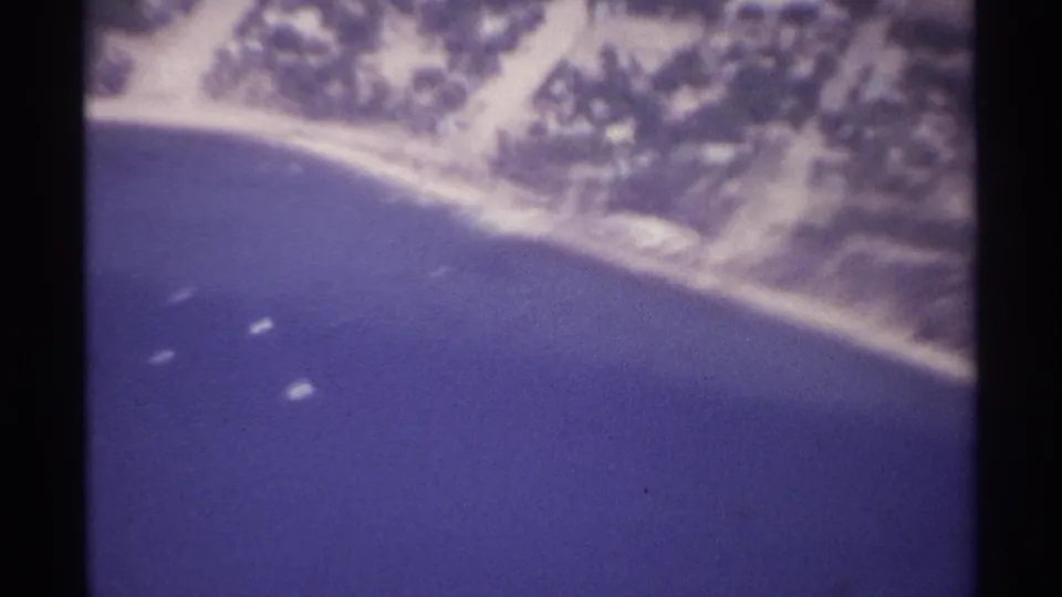 Soft 8mm aerial frame of the La Paz coastline in 1974, with open water, shoreline, and scattered boats below.