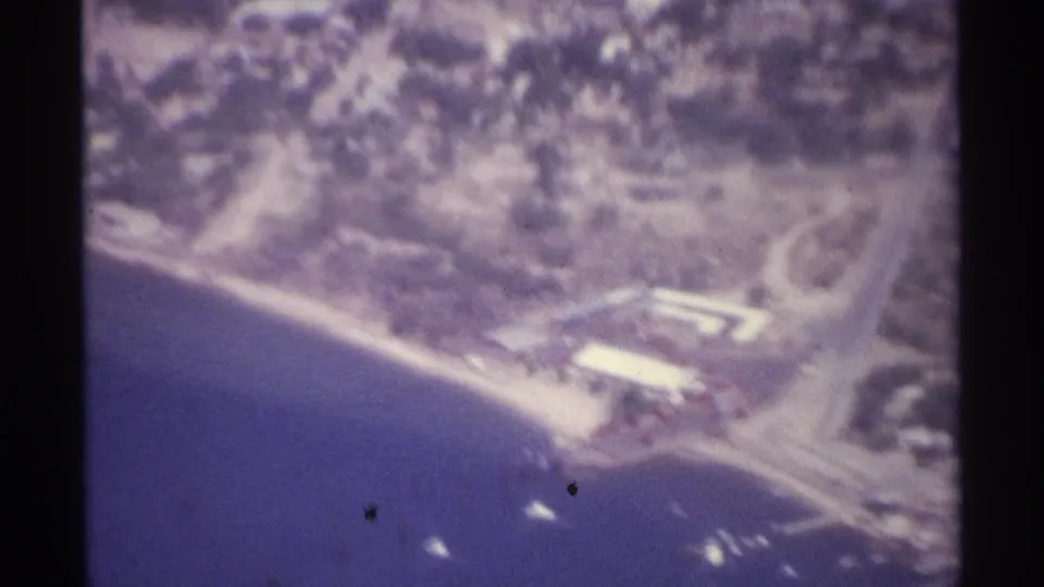 1974 La Paz, Mexico aerial frame showing a waterfront area, white-roofed structure, roads, shoreline, and boats in the bay.