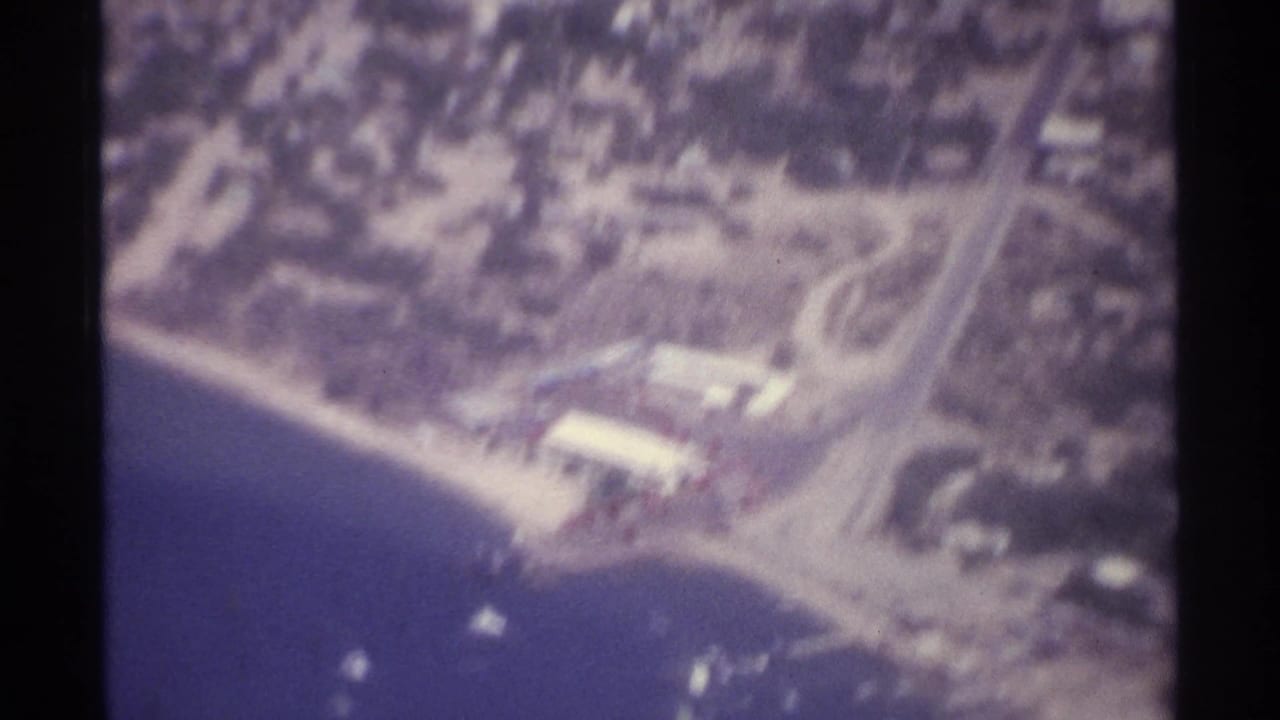 Soft aerial view of shoreline, water, roads, and scattered buildings.