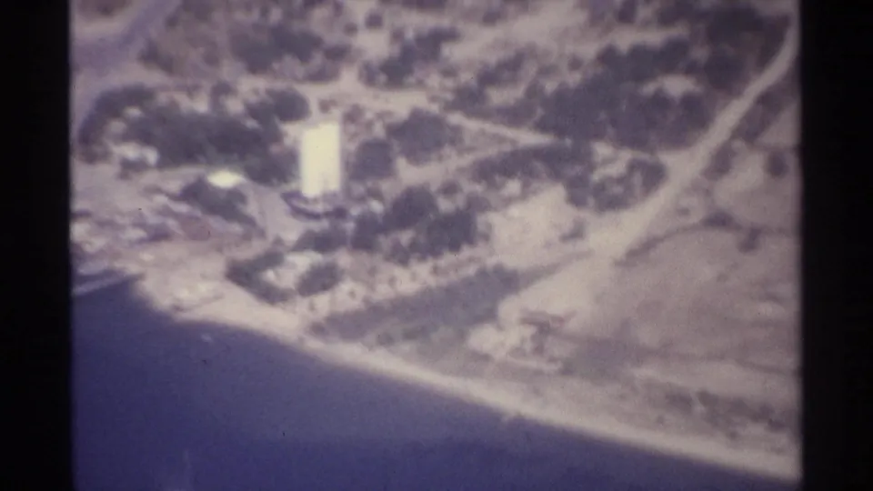 1974 La Paz aerial 8mm frame with shoreline water, pale beach, curving roads, and a bright rectangular building.
