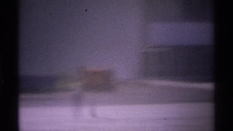 Blurry 1974 La Paz travel frame with a flagpole at right, muted sky, and soft ground or vehicle shapes below.