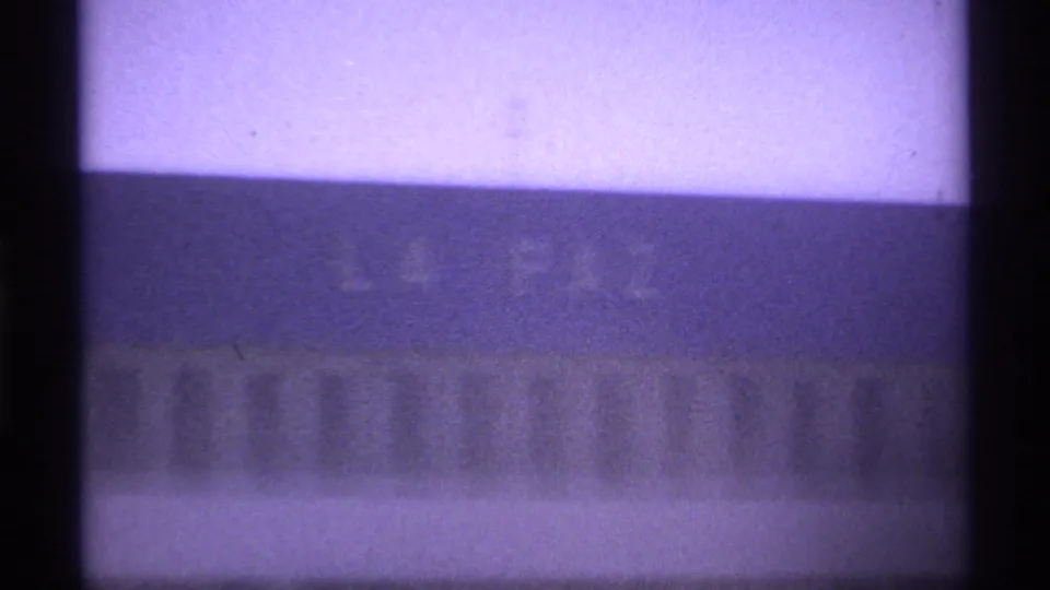 Soft 1974 La Paz frame showing a pale building band and indistinct lettering, with heavy grain and vertical foreground bars.