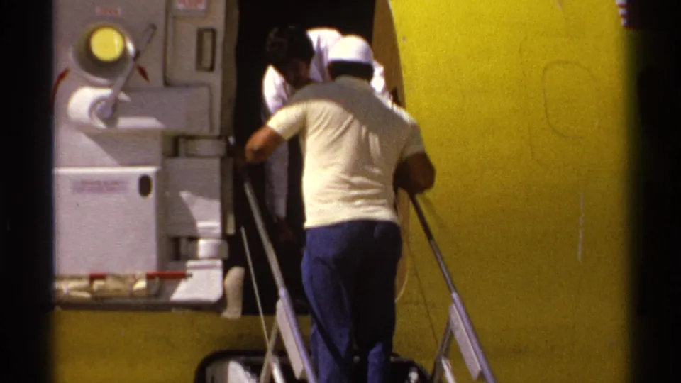 1974 La Paz airport frame showing a man in a light shirt guiding passengers down aircraft stairs beside a yellow commercial aircraft.