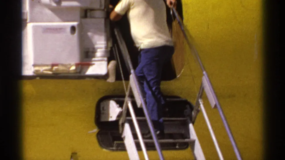 1974 La Paz home-movie frame showing a man boarding a yellow aircraft by metal stairs on the airport tarmac.