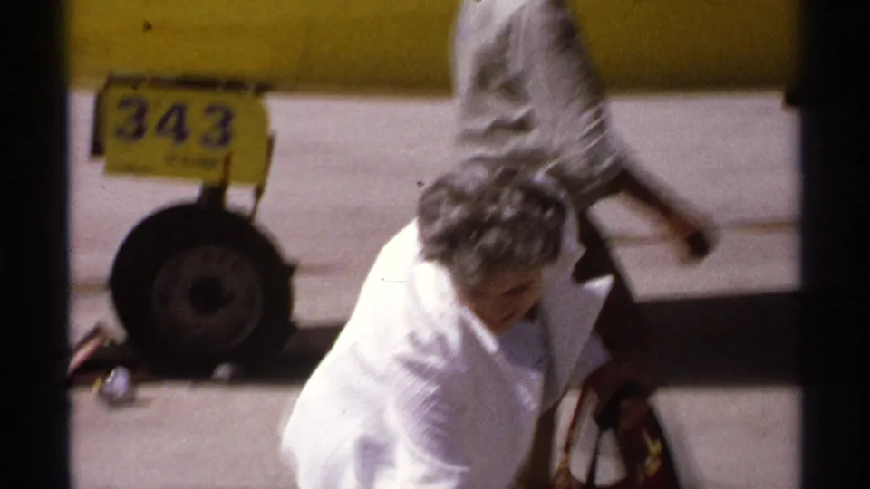 1974 La Paz frame showing a woman in white near luggage and landing gear beside the yellow aircraft on the tarmac.