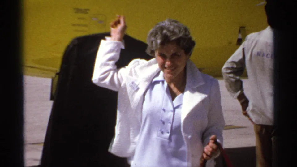 1974 La Paz home-movie frame of a woman in a white jacket walking on the tarmac beside the yellow aircraft and other travelers.
