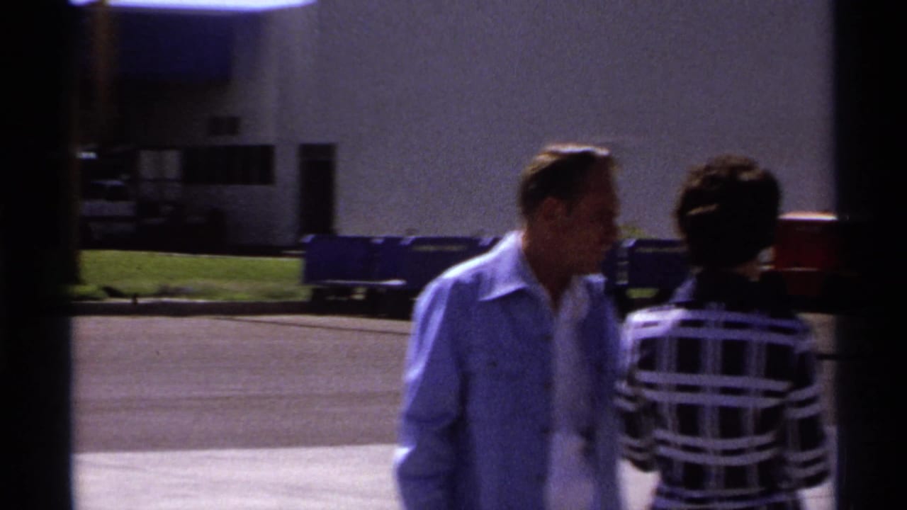 In La Paz, Mexico in 1974, a man in a light blue shirt walks beside a woman in a black-and-white plaid dress outside a pale building.