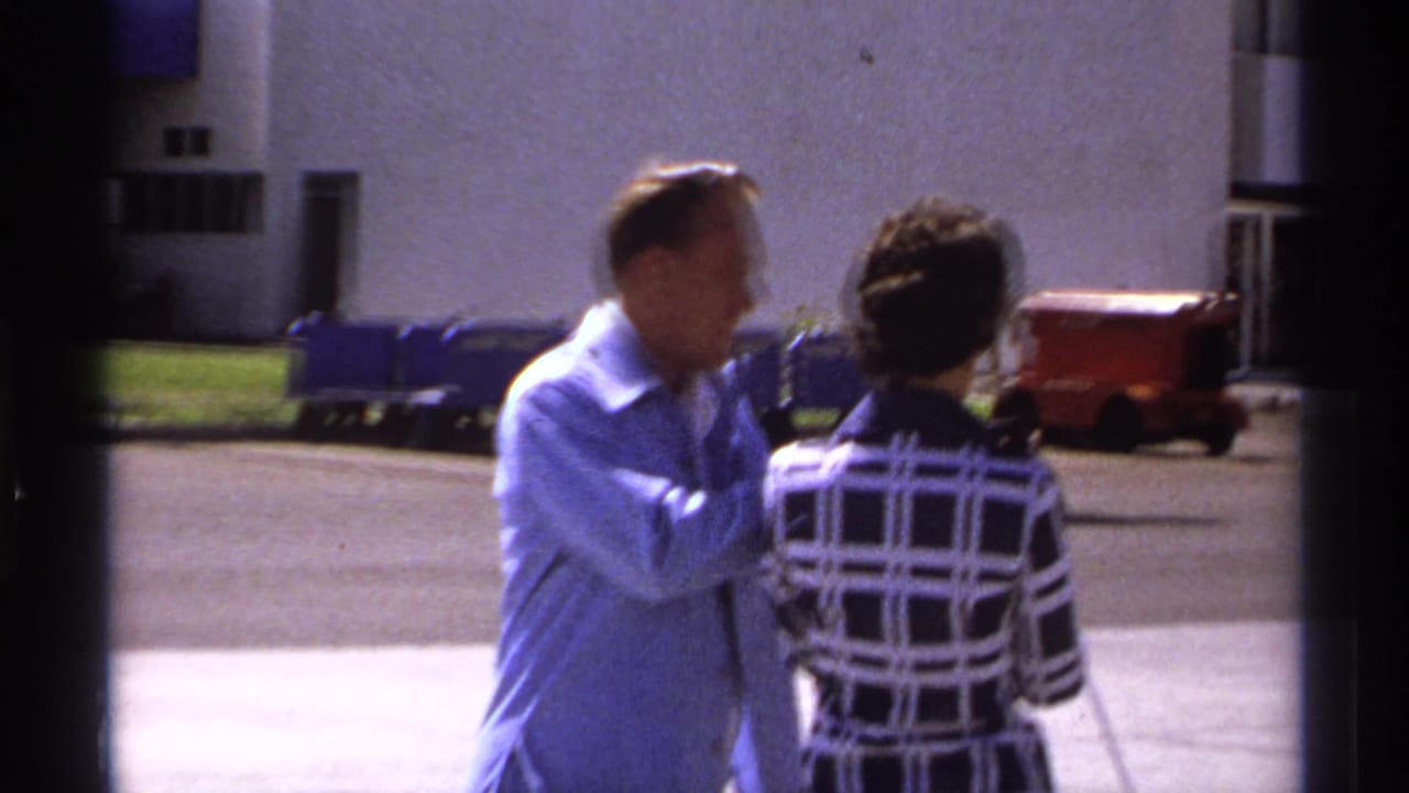 Man and woman continue walking near pavement and a pale building.