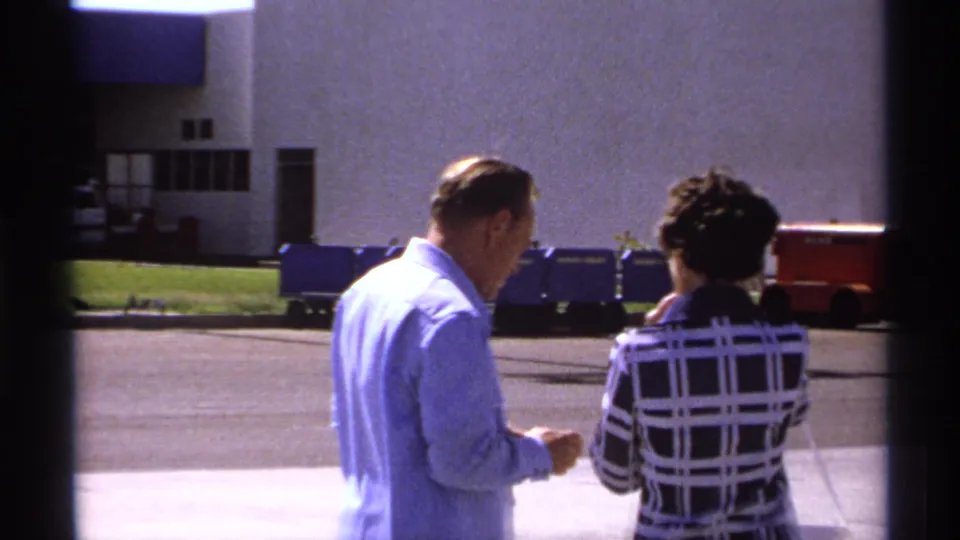 The man appears to check a small paper while the woman in the plaid dress walks beside him in the sunny exterior setting.