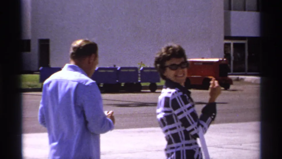 The woman in sunglasses turns toward the camera and smiles while holding a small paper, with the man and building exterior nearby.