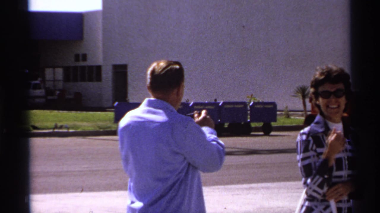 Man in a light blue shirt gestures toward a smiling woman outside a modern building.