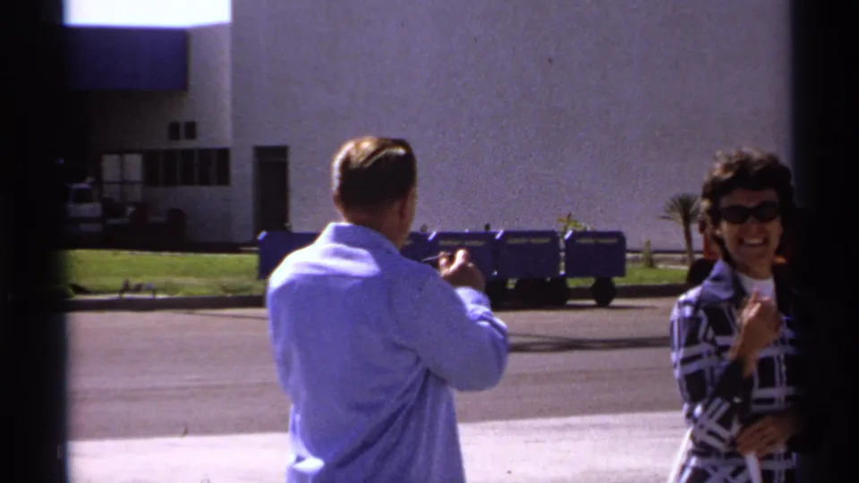 1974 La Paz home-movie frame showing a man gesturing while a woman in sunglasses smiles near a modern building facade.