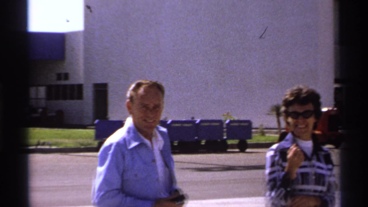 Man and smiling woman stand outside with blue carts and a building behind them.