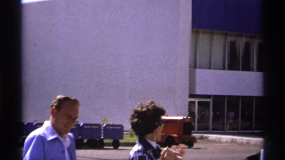 1974 La Paz frame with two adults outside a modern building, blue carts, and a red vehicle in the background.