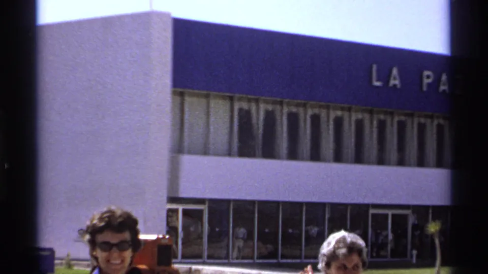 A smiling woman in sunglasses stands before a blue-and-white building facade in 1974 La Paz home-movie footage.