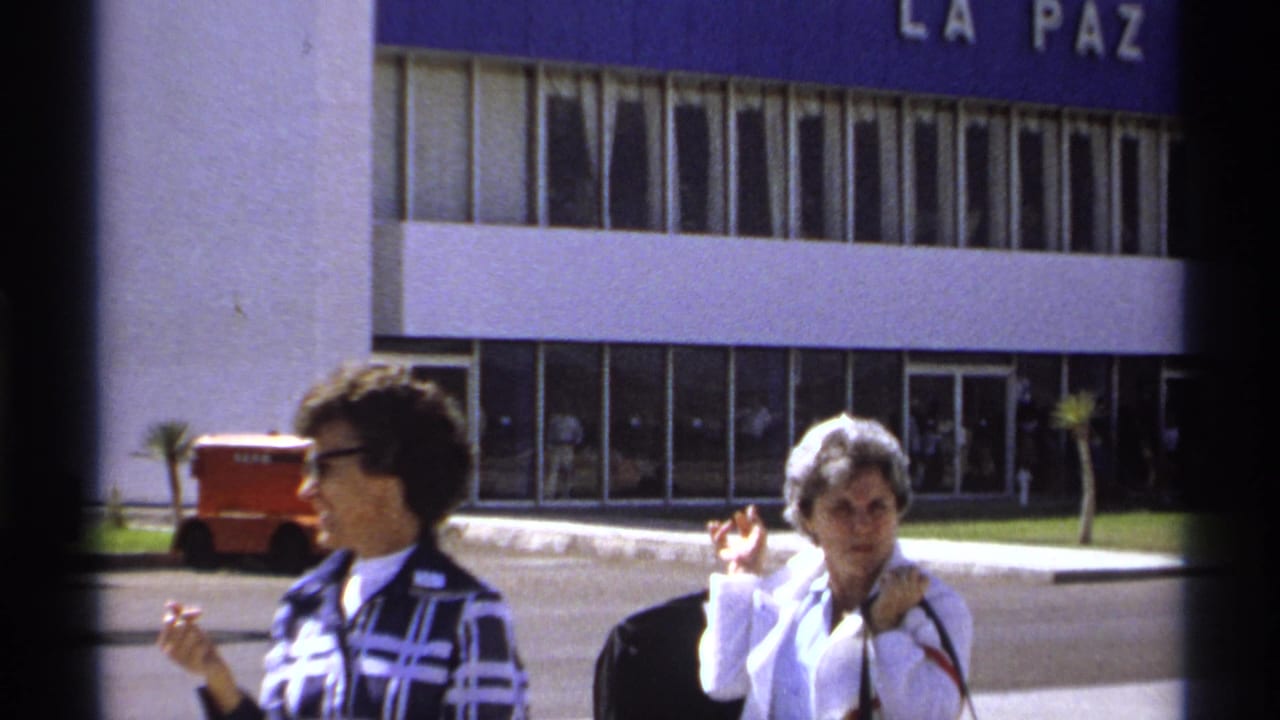 Two adult women smile and gesture outside a La Paz airport building in 1974 home-movie footage.