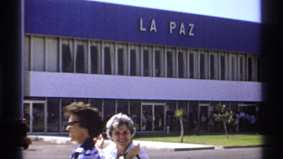 Two adult women stand near the La Paz airport entrance while people gather near the terminal doors.