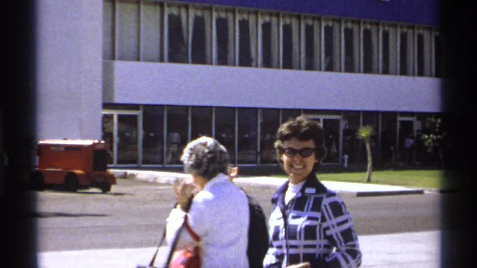 A woman in sunglasses smiles toward the camera outside the La Paz airport while another traveler turns away.