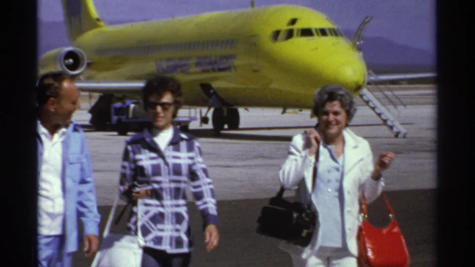 A closer 1974 home-movie frame shows three adult travelers on the La Paz tarmac beside a bright yellow jet.