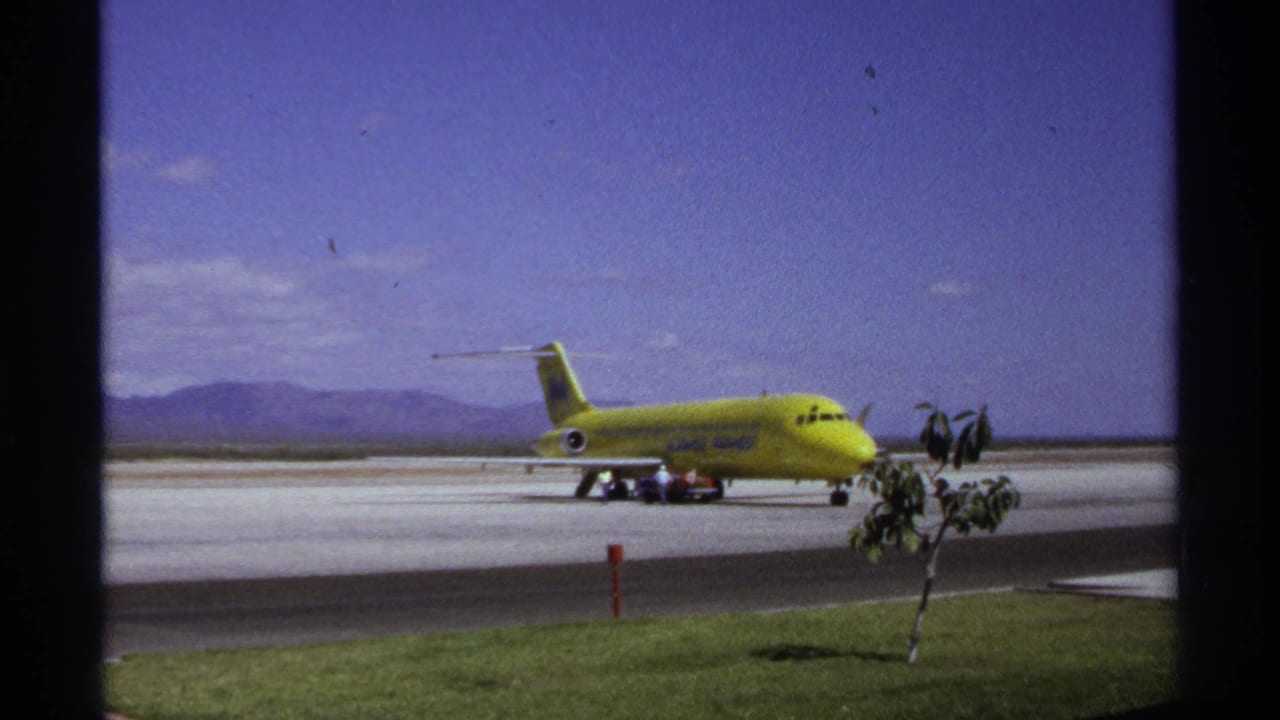Yellow T-tail airliner parked on a runway with service vehicles, small figures, trees, grass, and mountains.