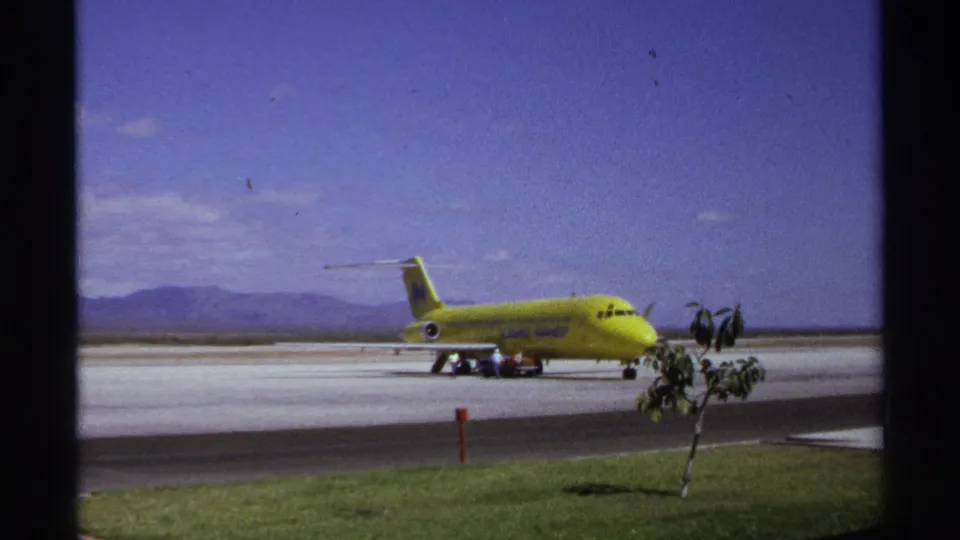 1974 La Paz, Mexico airport view of a yellow commercial jet on the tarmac with service activity and distant mountains.