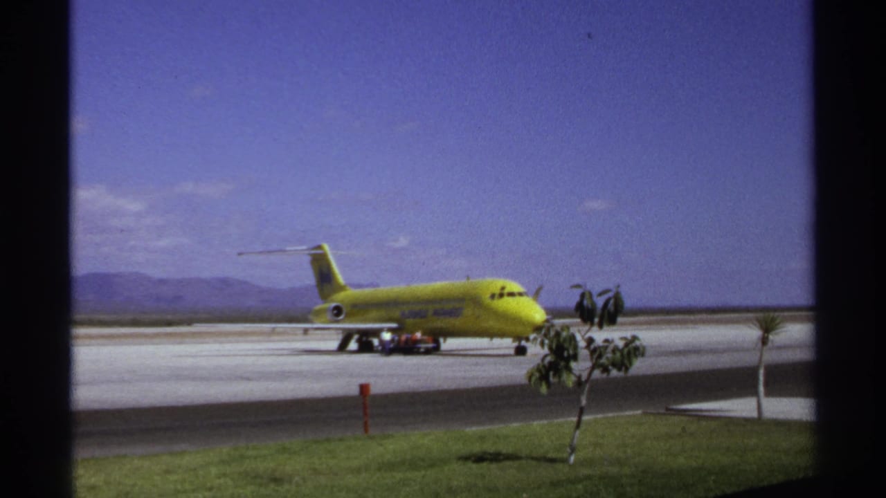 Yellow jet aircraft parked beside runway pavement with ground activity, foreground trees, blue sky, and mountains.
