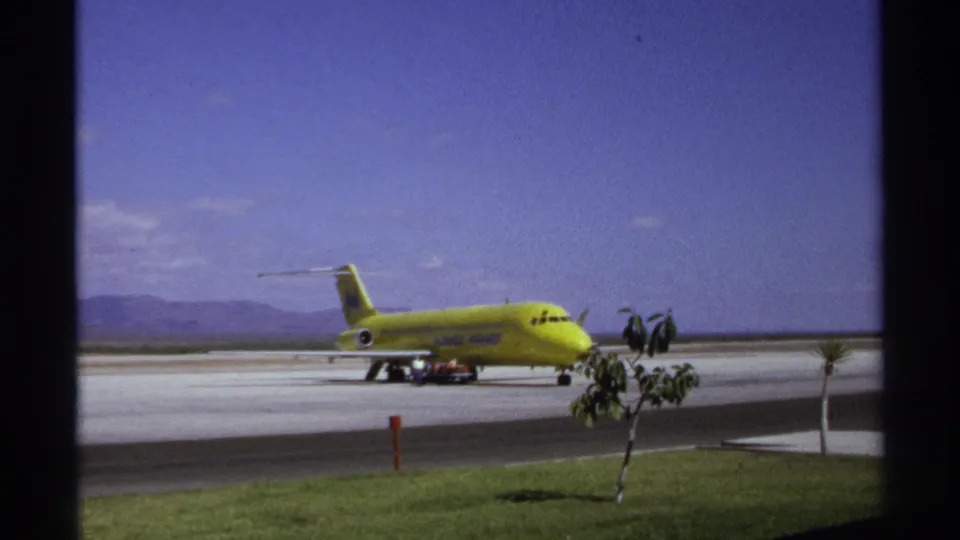 A yellow T-tail jet at the La Paz airport in 1974, seen from a visitor viewpoint across grass and runway pavement.