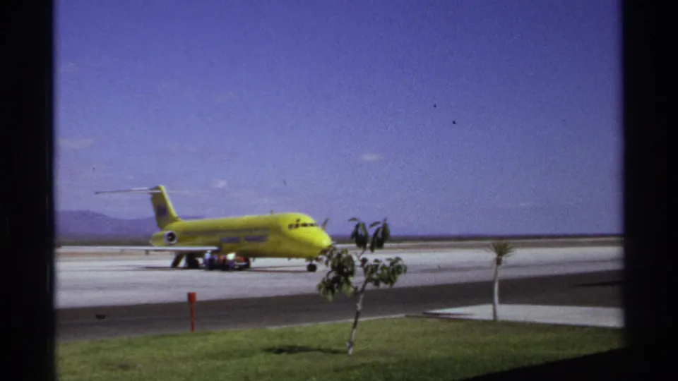 La Paz airport tarmac scene with a yellow airliner shifted left in frame, foreground grass, small trees, and 8mm film grain.