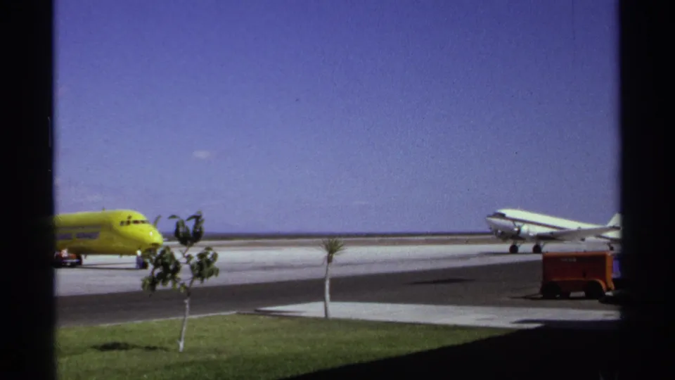 1974 La Paz airport scene with a yellow aircraft, a white propeller plane, small palms, grass, and a red ground cart at the edge.