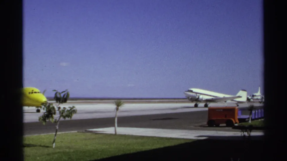 1974 La Paz tarmac view with the yellow plane partly leaving frame, a white propeller aircraft, palms, grass, and red ground equipment.